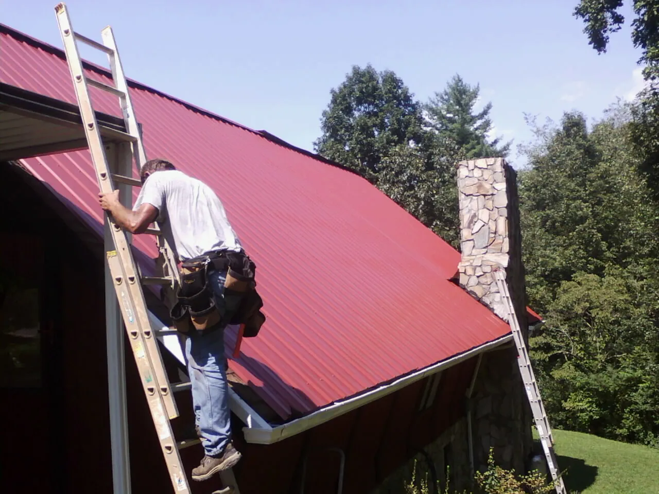 Roofer on a ladder beside a red metal roof and stone chimney — Soffit & Fascia Repair by Roofing Brothers in Hallsboro